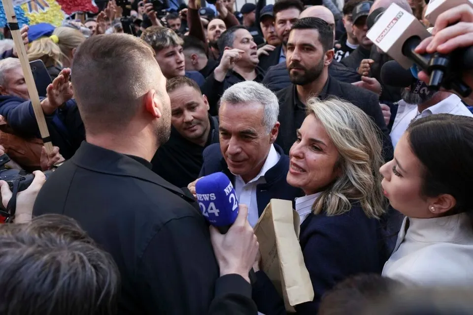 O femeie cu părul blond și un bărbat cu părul cărunt sunt înconjurați de o mulțime de oameni și microfoane.