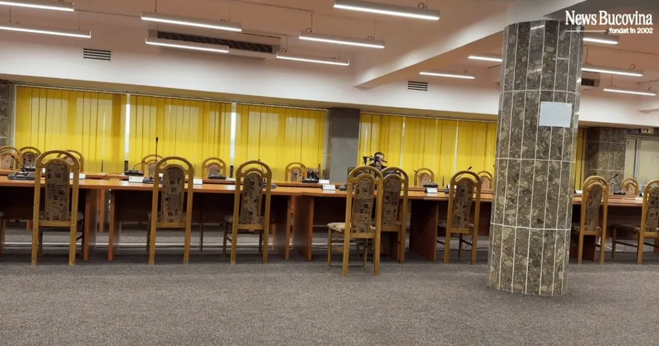 Un rând de scaune de lemn cu spătare înalte este așezat în fața unor mese lungi într-o încăpere cu pardoseală gri și draperii galbene.