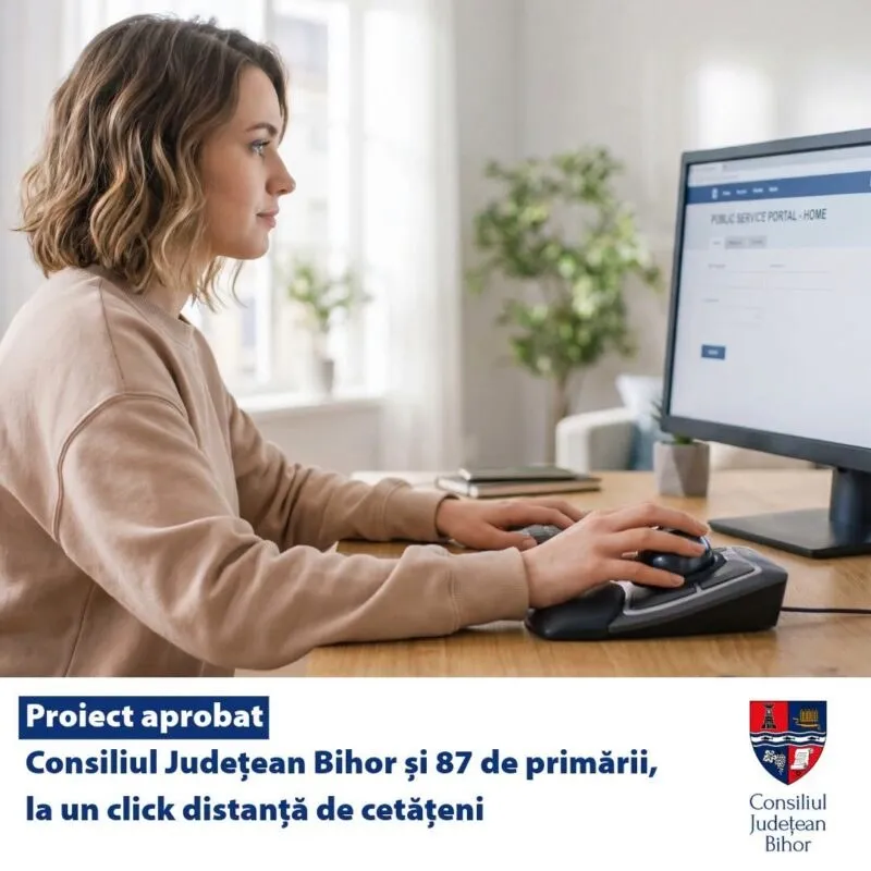 O femeie cu părul scurt și blond stă la o masă de lemn și folosește un mouse lângă un monitor de calculator.
