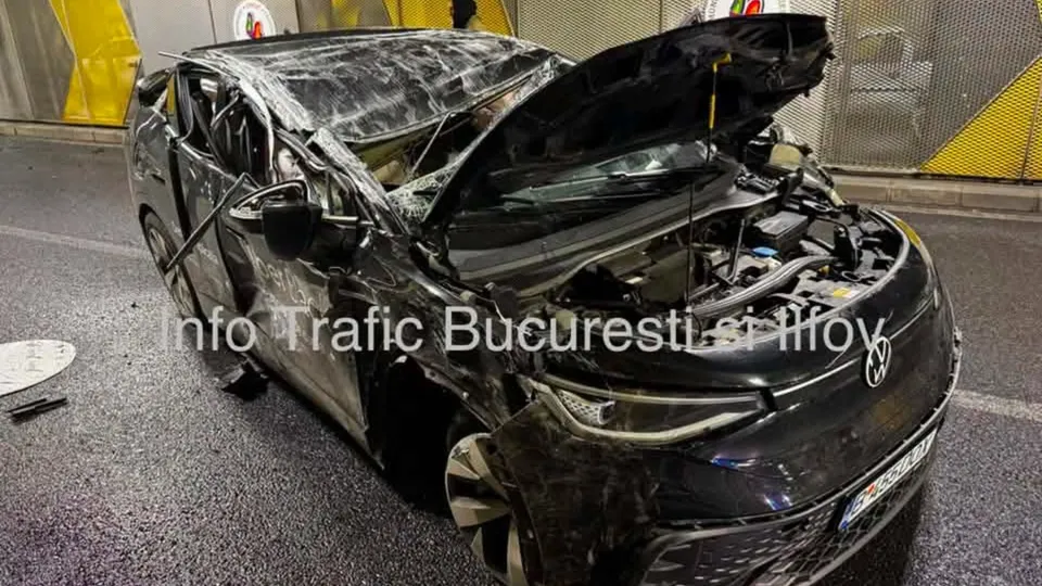 Un autovehicul negru cu capota ridicată și partea frontală avariată stă pe un drum asfaltat.