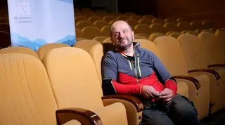 Un bărbat cu barbă și haină sportivă gri și roșie stă așezat pe un scaun de cinema galben într-o sală de proiecție.