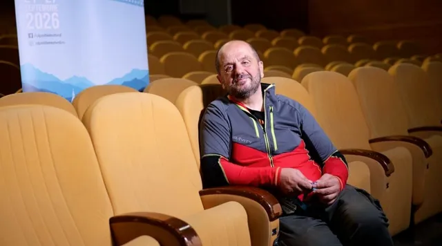 Un bărbat cu barbă și haină sportivă gri și roșie stă așezat pe un scaun de cinema galben într-o sală de proiecție.