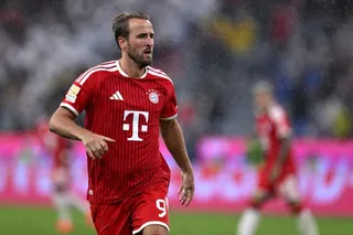 Un fotbalist cu barbă poartă un echipament sportiv roșu cu detalii albe și un logo alb pe piept, fiind surprins pe un teren de fotbal.