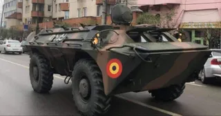 Un vehicul militar camuflat cu roți mari circulă pe un drum asfaltat lângă clădiri urbane și mașini albe.