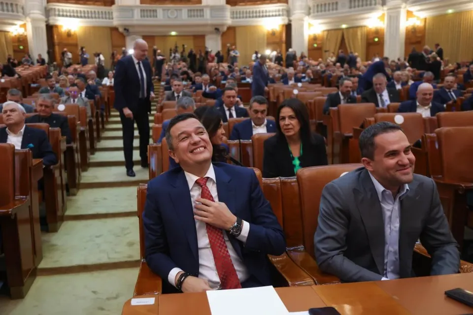 Un grup de bărbați în costume și sacouri stau așezați în scaune de lemn într-o sală de conferințe cu mulți oameni în fundal.