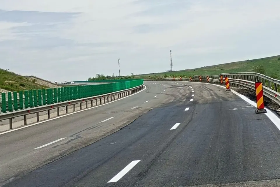 O autostradă cu asfalt negru și linii albe marcate curbează spre stânga, fiind delimitată de un parapet metalic și bariere verzi pe partea stângă.