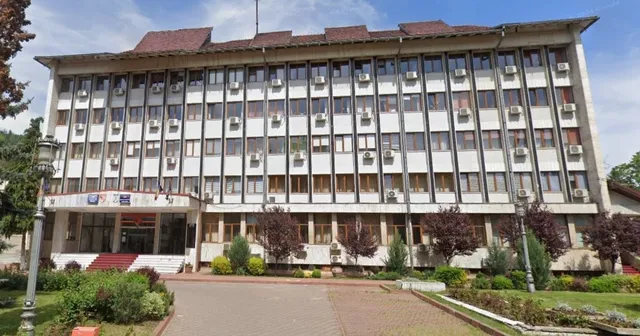 O clădire cu mai multe etaje prezintă numeroase ferestre și unități de aer condiționat montate pe fațadă, fiind înconjurată de vegetație și un drum.