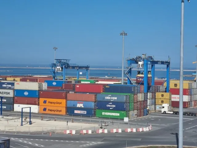 Containerul de marfă este stivuit pe un port în prezența unor macarale portuare sub un cer senin.