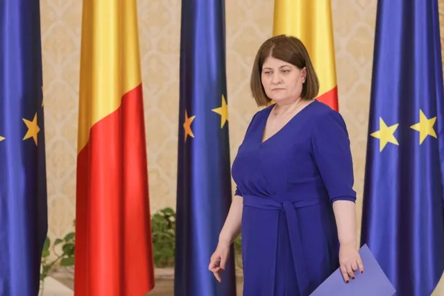 O femeie cu părul scurt și brunet purtând o rochie albastră stă în fața mai multor steaguri albastre cu stele galbene și unui steag tricolor.