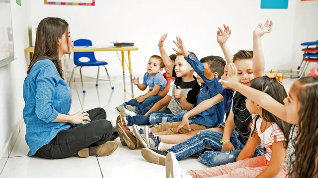 O femeie cu o cămașă albastră stă pe podea lângă un grup de copii care stau așezați pe podea și ridică mâinile.