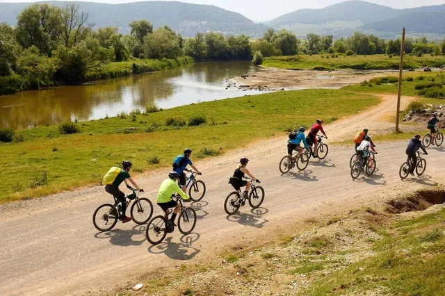 Oamenii merg pe biciclete pe un drum de pământ lângă un râu, într-un peisaj verde cu dealuri în fundal.