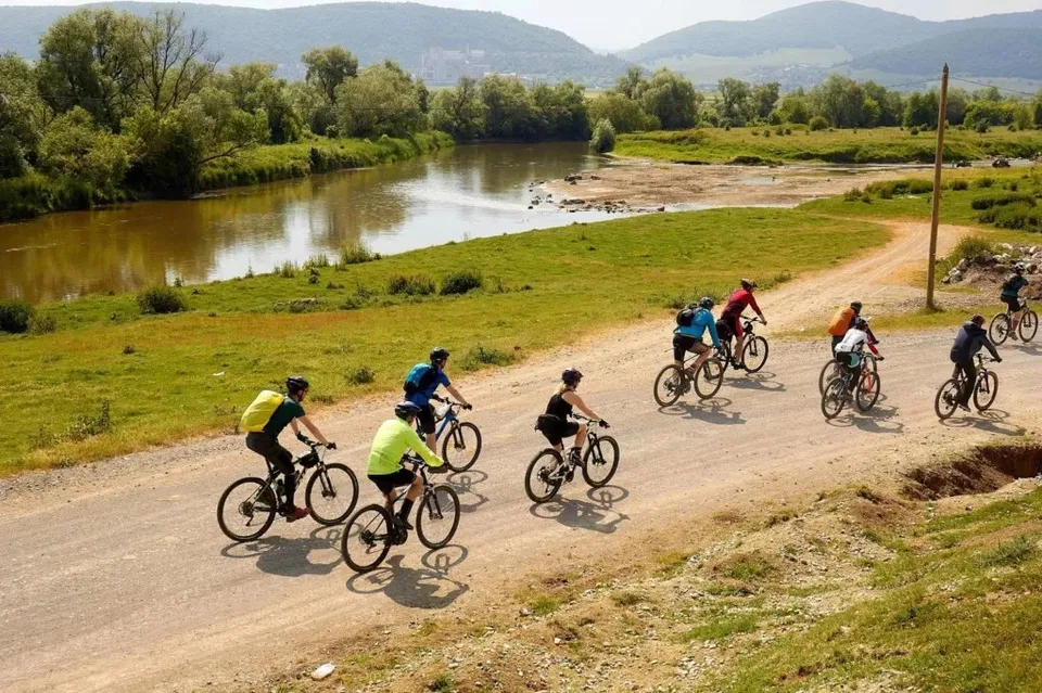 Oamenii merg pe biciclete pe un drum de pământ lângă un râu, într-un peisaj verde cu dealuri în fundal.