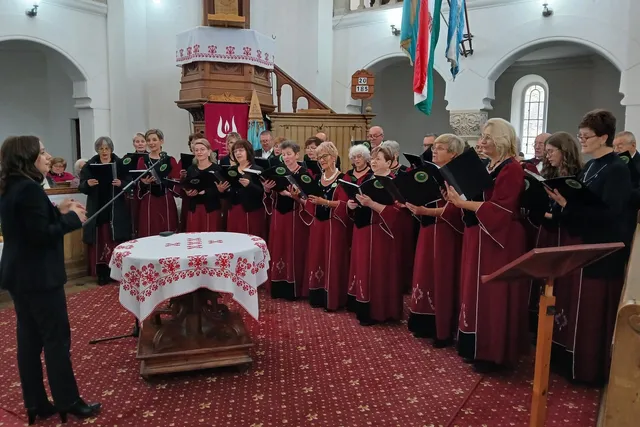 Un grup de femei îmbrăcate în rochii roșii și negre cântă ținând dosare negre într-o încăpere cu podea cu covor roșu și un pupitru.