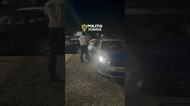 Un vehicul de poliție cu luminile aprinse este parcat pe un teren pietruios lângă un om care poartă o cămașă deschisă și pantaloni în noapte.