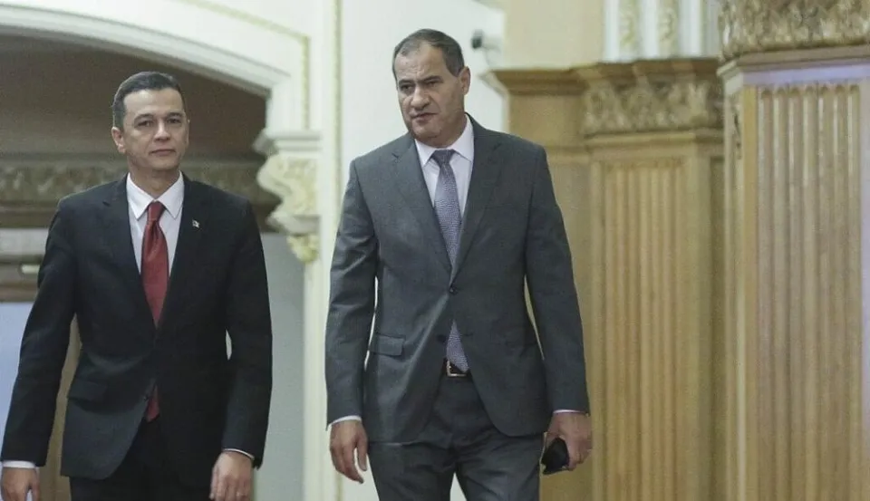 Doi bărbați în costume și cravate merg printr-un hol cu coloane decorate și elemente arhitecturale clasice.