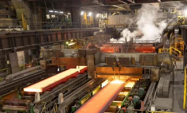 O linie de producție industrială prezintă role de metal incandescente pe o bandă transportoare într-un spațiu mare cu structuri metalice și abur.