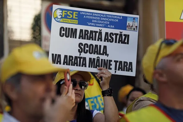 O persoană ține un panou alb cu text negru, în timp ce alți oameni cu căciuli galbene stau în apropiere.