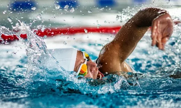 Un înotător cu căciulă albă și ochelari de înot colorați înoată într-o piscină, provocând stropi de apă în jurul capului și brațului său.