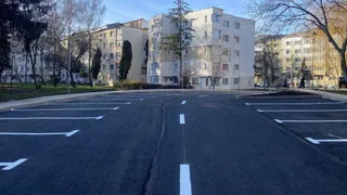 O parcare proaspăt asfaltată cu locuri marcate cu linii albe, în fața unor blocuri de locuințe într-un cartier urban.