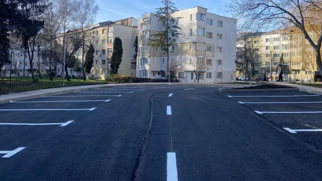O parcare proaspăt asfaltată cu locuri marcate cu linii albe, în fața unor blocuri de locuințe într-un cartier urban.