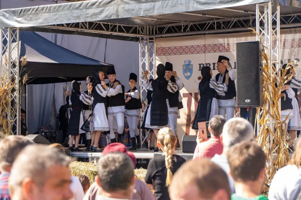 Un grup de copii îmbrăcați în costume tradiționale albe și negre dansează pe o scenă în fața unui public.
