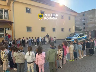 Un grup de copii stă în rând pe o curte pavată lângă o mașină de poliție și o clădire galbenă sub lumina soarelui.