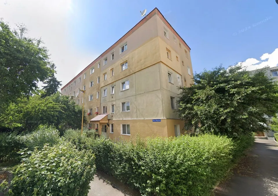 Un bloc de apartamente cu fațadă de două culori este înconjurat de vegetație și copaci sub un cer senin.