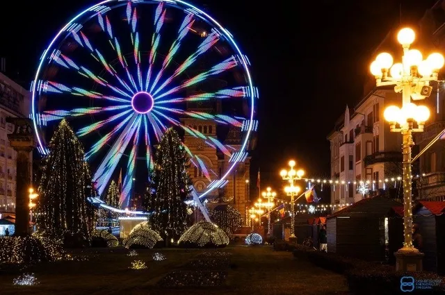 O roată feroviară iluminată cu lumini colorate strălucește pe cerul de noapte lângă copaci decorați și felinare de stradă cu globuri luminoase.