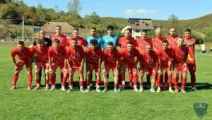 O echipă de fotbaliști în echipamente roșii și albe pozează împreună pe un teren de fotbal verde sub un cer senin.