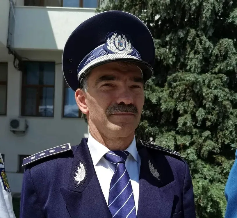 Un bărbat cu mustață poartă uniformă de polițist, pălărie cu emblemă și cravată albastră cu dungi diagonale, stând în fața unei clădiri și a unui copac.
