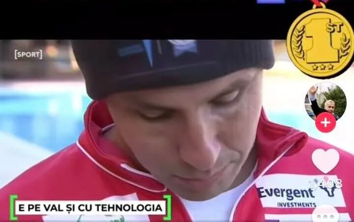Un bărbat cu o șapcă neagră și o jachetă roșie cu detalii albe privește în jos în fața unei ape albastre.