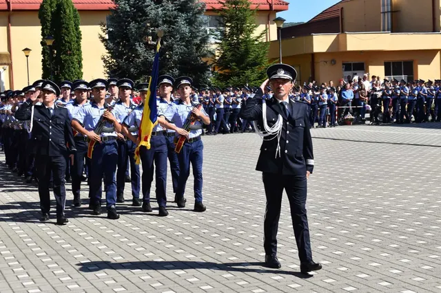 Un ofițer în uniformă neagră salută în prim-plan, în timp ce un grup de agenți în uniformă albastră stă în rânduri pe o curte pavată.