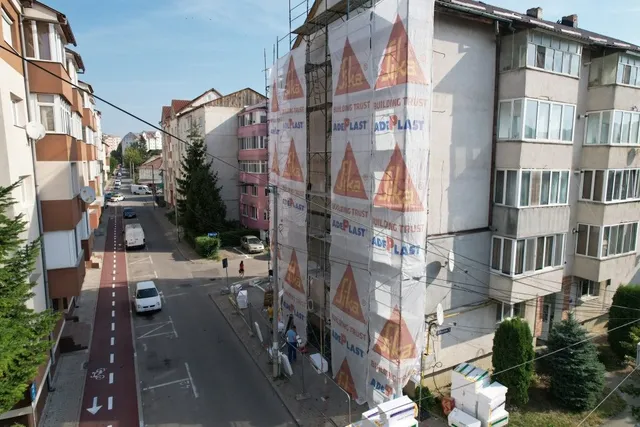 Un bloc de locuințe este acoperit de un panou de protecție alb cu logo-uri portocalii, situat lângă o stradă cu mașini și vegetație.