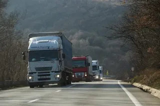 Mai multe camioane circulă pe un drum asfaltat lângă un versant de deal împădurit sub un cer înnorat.