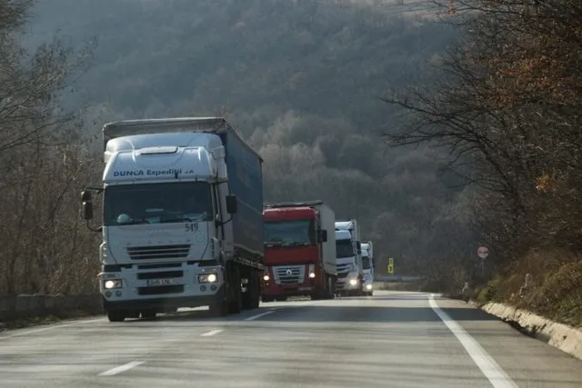 Mai multe camioane circulă pe un drum asfaltat lângă un versant de deal împădurit sub un cer înnorat.