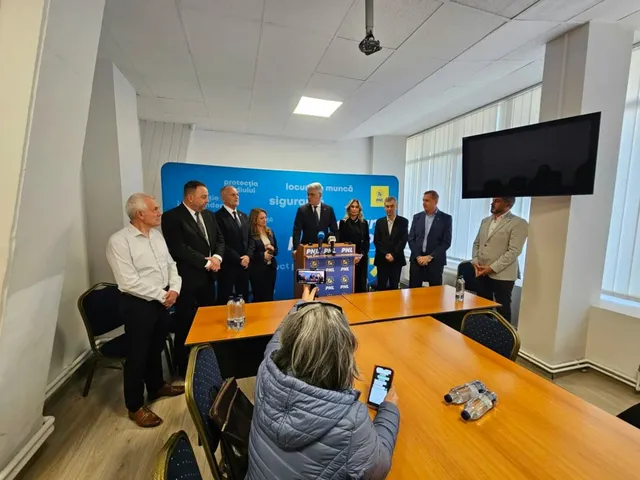 Un grup de oameni stă în picioare în fața unui panou albastru într-o încăpere cu mese de lemn și un ecran negru suspendat.