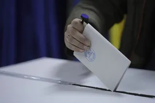 O mână ține un plic alb și îl introduce într-o deschidere pe o suprafață plană, în fața unui fundal albastru.