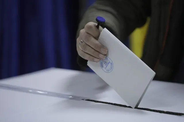 O mână ține un plic alb și îl introduce într-o deschidere pe o suprafață plană, în fața unui fundal albastru.
