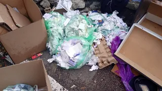 Mai multe saci de plastic verzi și albi conțin diverse obiecte de hârtie și resturi de carton pe un trotuar lângă cutii deschise.