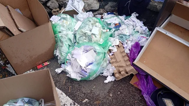 Mai multe saci de plastic verzi și albi conțin diverse obiecte de hârtie și resturi de carton pe un trotuar lângă cutii deschise.