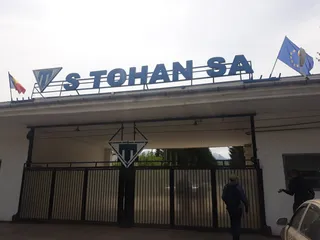 Un semn albastru cu textul S TOHAN SA este montat pe acoperișul unei clădiri albe, lângă două steaguri.