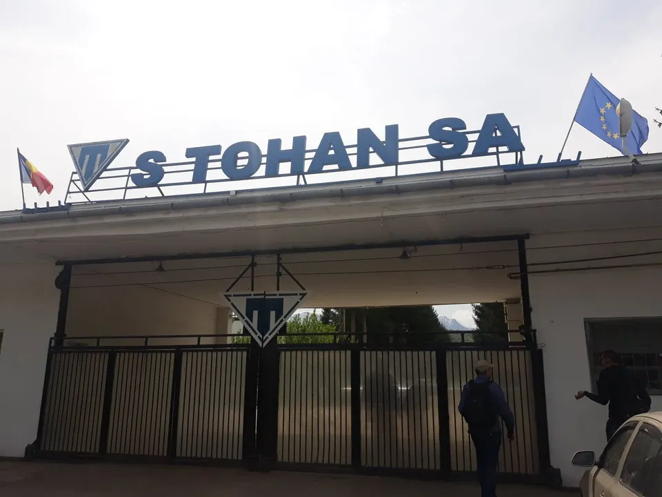 Un semn albastru cu textul S TOHAN SA este montat pe acoperișul unei clădiri albe, lângă două steaguri.
