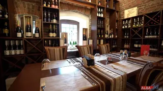 Mesele de lemn sunt pregătite cu seturi de tacâmuri și fețe de masă în fața unor rafturi de lemn pline cu sticle de vin.