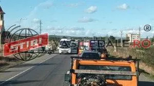 O șosea cu vehicule oprite, un autobuz alb, autoturisme, o platformă portocalie și echipaje în fundal.