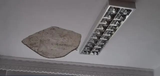 O bucată de tencuială este desprinsă de la un tavan alb lângă un corp de iluminat rectangular încastrat.