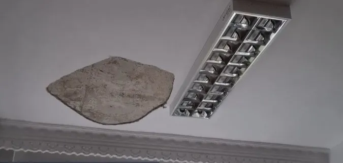 O bucată de tencuială este desprinsă de la un tavan alb lângă un corp de iluminat rectangular încastrat.