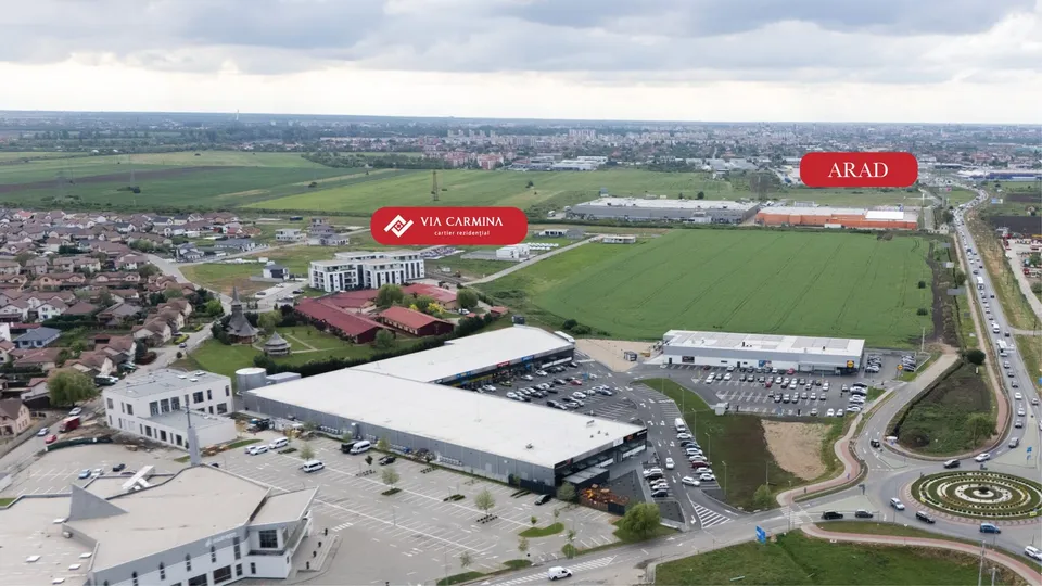 O vedere aeriană prezintă clădiri comerciale, terenuri verzi, parcuri de staționare cu mașini și o stradă cu trafic lângă un sens giratoriu.