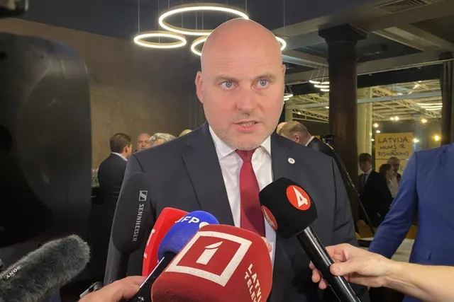 Un bărbat cu capul chel, purtând costum închis și cravată roșie, este înconjurat de microfoane în fața unor persoane din fundal.