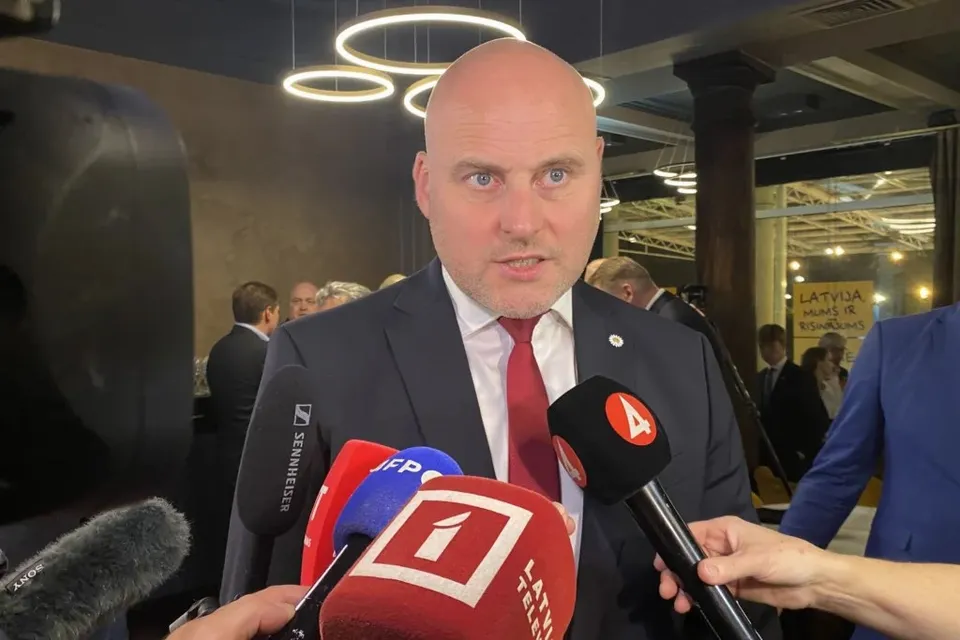 Un bărbat cu capul chel, purtând costum închis și cravată roșie, este înconjurat de microfoane în fața unor persoane din fundal.