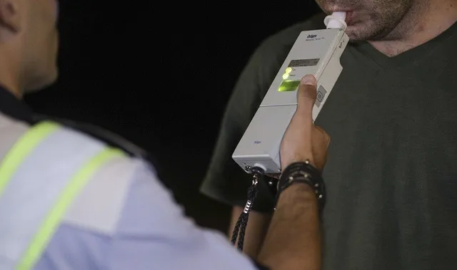 Un dispozitiv alb este ținut de o mână în fața gurii unui bărbat, în timp ce o persoană în uniformă stă în prim-plan.